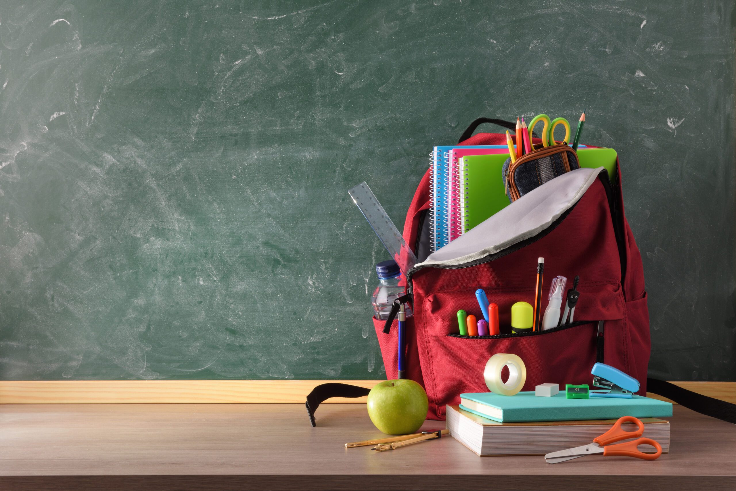 Backpack full of school supplies on wood table and green blackboard with blank space for writing front view